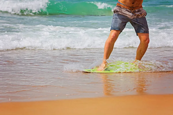 Jovem esportivo homem está correndo atrás de seu skimboard amarelo e azul na praia em um dia ensolarado — Fotografia de Stock