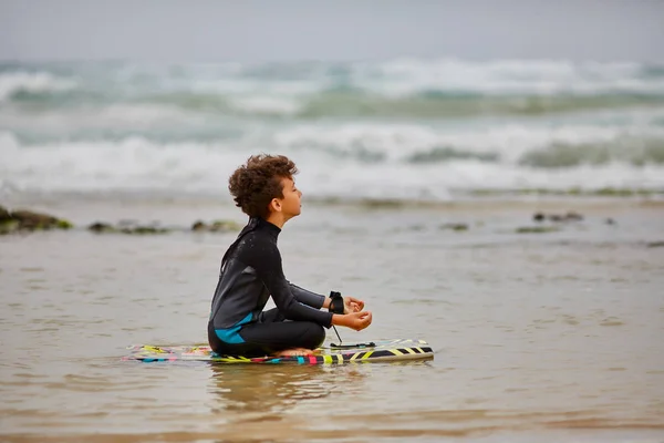 Kleiner Junge im Surferanzug sitzt auf einer Tafel und meditiert am Strand. — Stockfoto