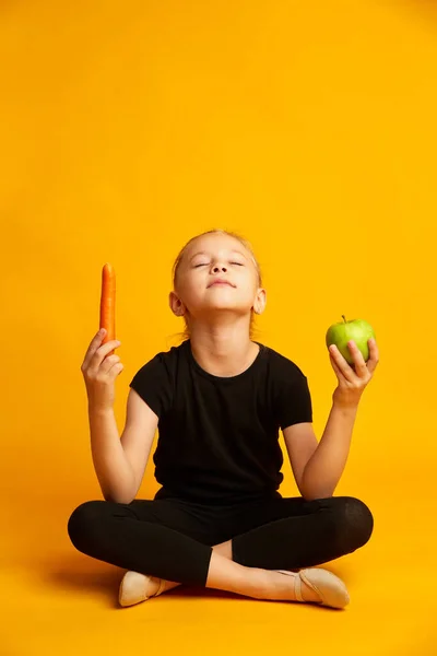 Atlética niña de siete años sosteniendo manzana verde y zanahoria en manos aisladas en amarillo —  Fotos de Stock