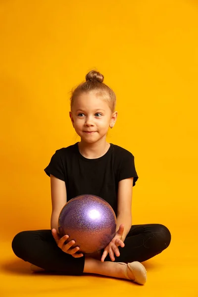 Estudio de tiro de la atractiva niña gimnasta rubia con leyendas negras y un traje de baño con una bola púrpura sentado piernas cruzadas contra el fondo amarillo — Foto de Stock