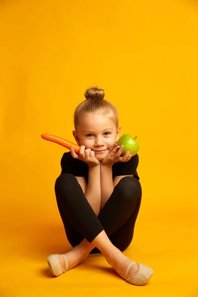 Pequeña deportista juguetona en ropa deportiva negra y zapatos Pointe sobre un fondo amarillo, sosteniendo una zanahoria y una manzana verde en sus manos — Foto de Stock