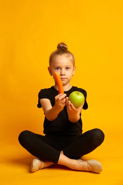 Niña aturdida en traje de baño y zapatos de baile, eligiendo comida saludable y mirando a la cámara, sentada con las piernas cruzadas sobre un fondo amarillo —  Fotos de Stock