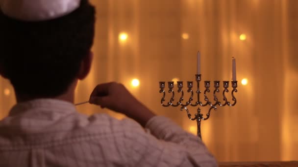 La vista desde la parte posterior de un niño enciende la primera vela de la menorá durante la fiesta judía de Hanukkah . — Vídeos de Stock