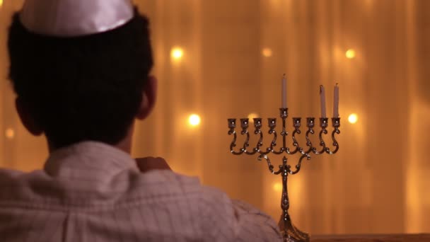 Visão traseira um menino judeu acende uma terceira vela menorah durante o feriado judaico de Hanukkah . — Vídeo de Stock