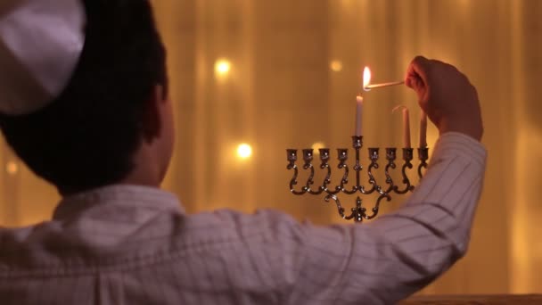 Zadní pohled na mladého chlapce rozsvítí druhou menorah svíčku během židovského svátku Chanuka — Stock video