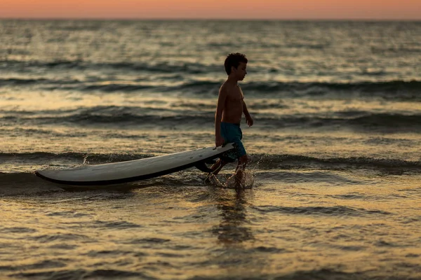 Silhueta de menino correndo ao longo da praia com uma prancha de surf durante o pôr do sol — Fotografia de Stock
