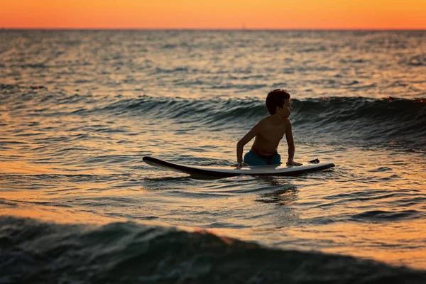Silhueta de um garotinho esperando por ondas ao pôr do sol — Fotografia de Stock