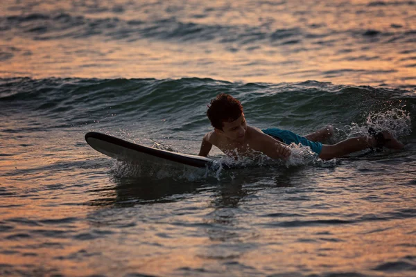 Menino surfista deitado em uma prancha de surf à noite na hora do pôr do sol — Fotografia de Stock