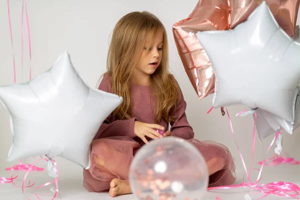 Happy brightful image of cute joyful little girl in tulle skirt sitting on present with balloons on white background — Stock Photo, Image