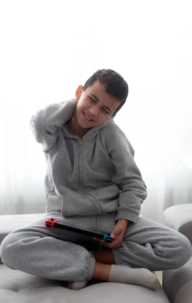 Schooljongen met een console in zijn handen lijdt aan rugpijn tijdens het zitten op een bank — Stockfoto