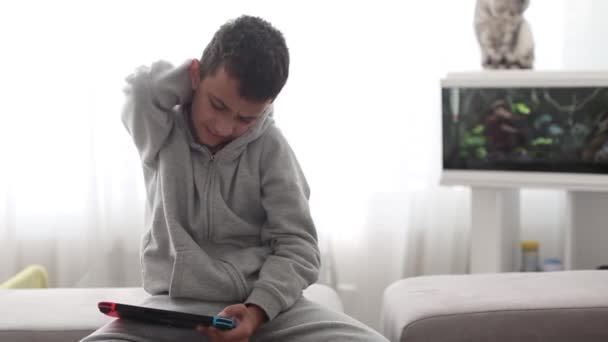 Estudante com um console em suas mãos sofre de dor nas costas enquanto sentado em um sofá — Vídeo de Stock