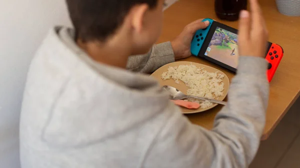 Kind eet rijst en surft op internet of speelt videospelletjes op de console. jongen eet slecht afgeleid door het spel. — Stockfoto