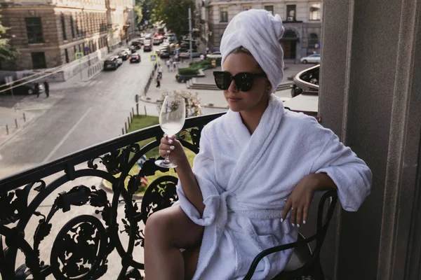 Giovane bella ragazza in vestaglia si siede su un balcone con un bicchiere di vino — Foto Stock
