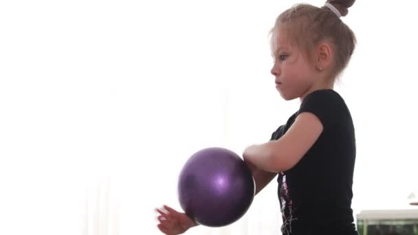 Menina de olhos azuis Toddler faz ginástica rítmica em um bodysuit preto, joga com uma bola em casa — Vídeo de Stock