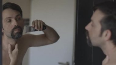 Genç beyaz adam sakalını elektrikli makasla kesiyor.