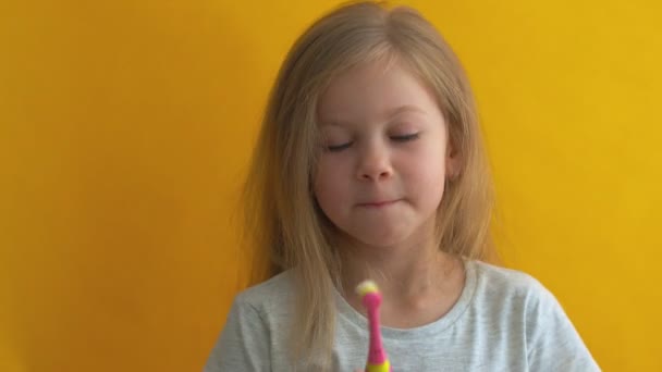 Обаятельная девчонка в серой футболке чистит зубы электрической детской зубной щеткой, глядя в камеру на желтом фоне — стоковое видео