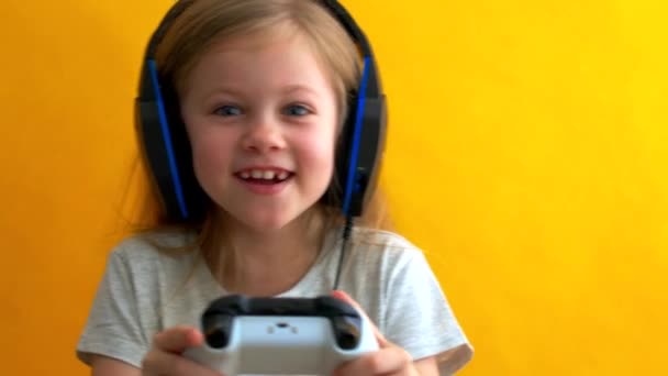 Pequena loira com olhos azuis em fones de ouvido joga um jogo de vídeo com um gamepad em um fundo amarelo — Vídeo de Stock