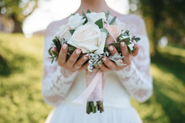 Gelin önünde bir buket beyaz gül tutuyor. Arka planda yeşillik var.