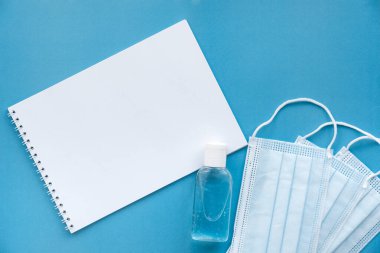 Üstten bakıldığında, mavi arka planda bir not defteri, tıbbi maskeler ve el dezenfektanı var. Defterde, mesajınız için boş bir sayfa.
