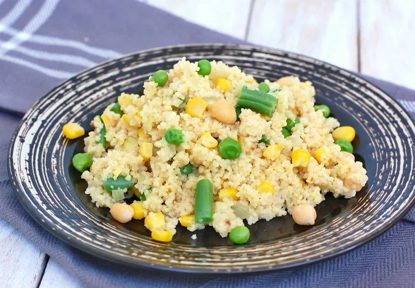 Healthy vegan meal with whole grain couscous, chickpeas, sweet corn, peas, green beans on dark plate on grey cloth on wooden background