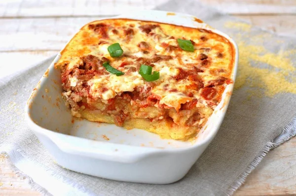 Polenta asada sin gluten con carne de cerdo, cebolla de primavera, especias en sartén blanca sobre tela gris sobre fondo de madera blanca —  Fotos de Stock