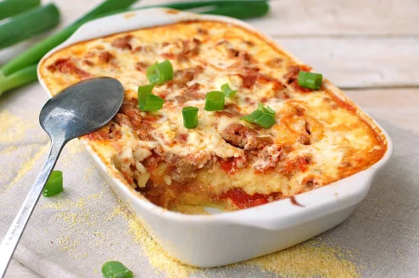 Polenta asada sin gluten con carne de cerdo, cebolla de primavera, especias en sartén blanca sobre tela gris sobre fondo de madera blanca —  Fotos de Stock