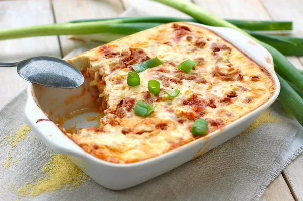 Polenta asada sin gluten con carne de cerdo, cebolla de primavera, especias en sartén blanca sobre tela gris sobre fondo de madera blanca —  Fotos de Stock