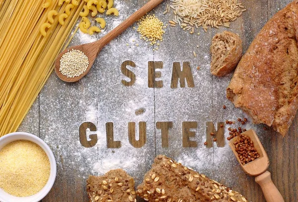 Gluten free flour and cereals millet, quinoa, corn flour polenta, brown buckwheat, basmati rice and pasta with text gluten free in Portuguese language with wooden spoon on brown wooden background