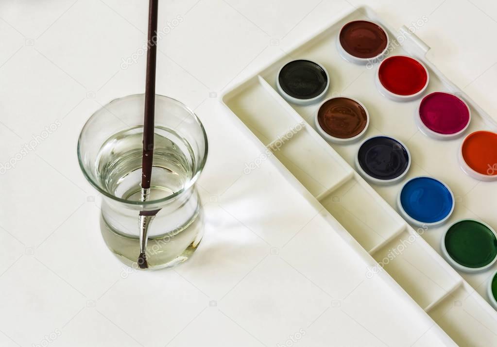 On a white surface is a set of watercolor colors and there is a glass with water and a brush