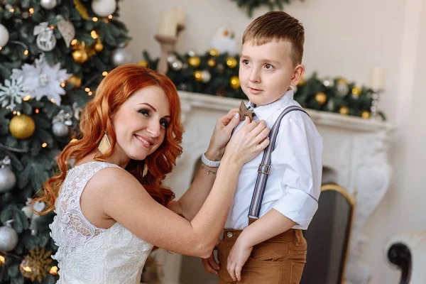 Joven madre viste a su hijo con una pajarita en el fondo de las decoraciones navideñas —  Fotos de Stock