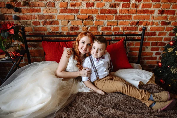 Navidad. Madre e hijo sentados en una cama en una habitación con una pared de ladrillo . —  Fotos de Stock