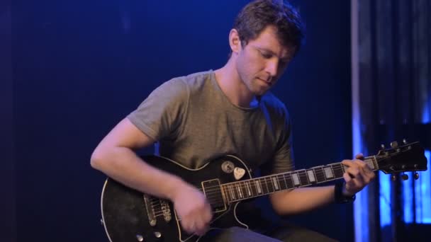 Мужчина сидит и играет на гитаре в темной комнате — стоковое видео