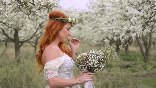 Red-haired beautiful woman in a luxurious dress and with a wreath on her head is standing in a flowering garden — Stock Video