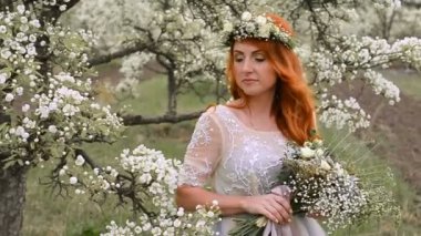 Kızıl saçlı güzel kadın lüks bir elbise ve başında bir çelenk ile çiçekli bahçe içinde duruyor