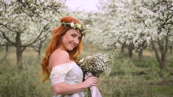 Red-haired beautiful woman in a luxurious dress and with a wreath on her head is standing in a flowering garden — Stock Video