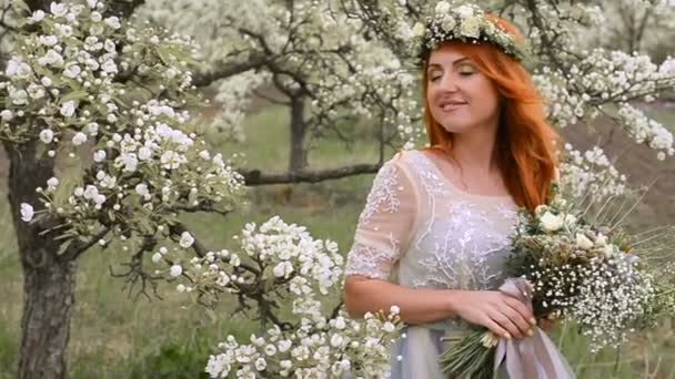 Red-haired beautiful woman in a luxurious dress and with a wreath on her head is standing in a flowering garden — Stock Video