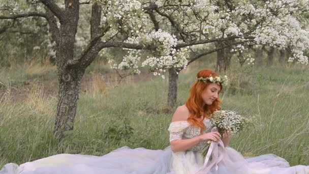 Red-haired woman dressed in a dress sits on the grass in a blooming garden — Stock Video