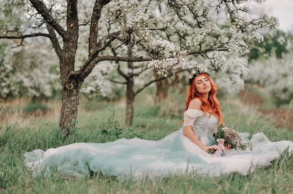 Jovem mulher em um vestido de luxo senta-se na grama em um jardim florescente — Fotografia de Stock