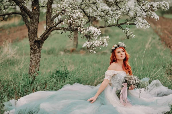 Young woman in a luxurious dress sits on the grass in a blooming garden — Stock Photo, Image