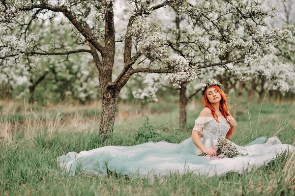 豪華なドレスを着た若い女性が咲く庭の芝生の上に座っています。 — ストック写真