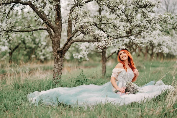 豪華なドレスを着た若い女性が咲く庭の芝生の上に座っています。 — ストック写真
