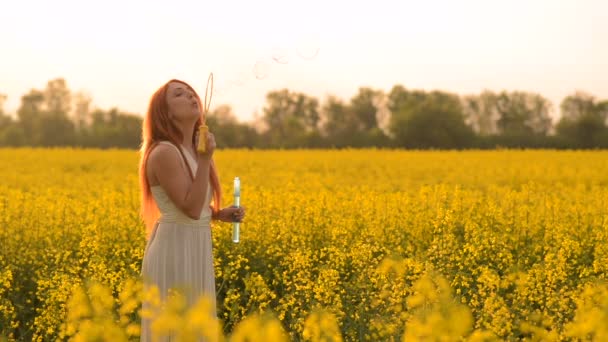 Young red-hair woman blowing bubbles at the camera outdoors in summer meadow — Stock Video