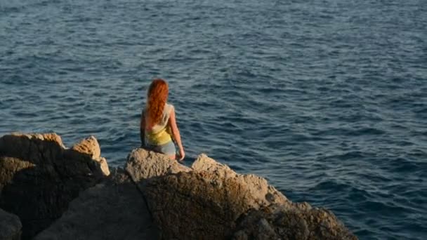 Молодая женщина с летящими волосами сидит на скалистом берегу моря — стоковое видео