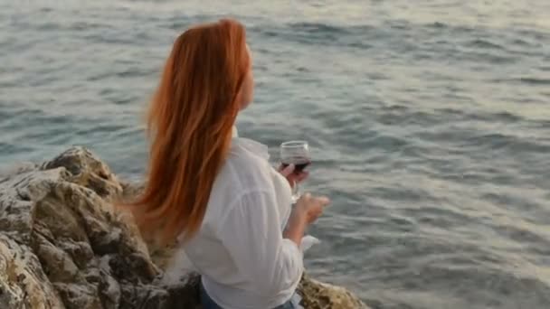 Giovane donna con i capelli volanti in piedi con un bicchiere di vino sulla spiaggia rocciosa del Mare Adriatico — Video Stock