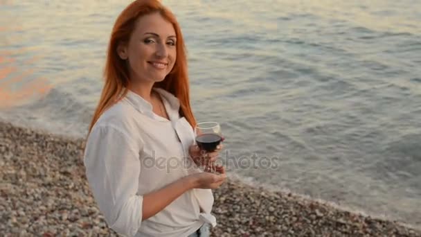 Junge Frau mit fliegendem Haar steht mit einem Glas Wein am felsigen Strand der Adria — Stockvideo