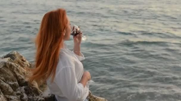 Mujer joven con el pelo volador de pie con una copa de vino en la playa rocosa del mar Adriático — Vídeos de Stock