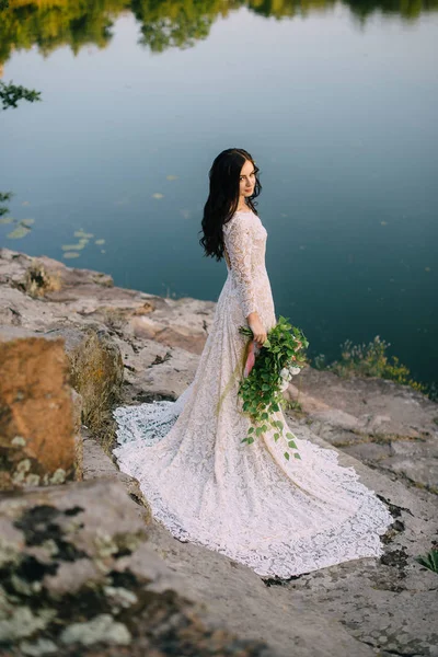 Giovane sposa in piedi sulla riva rocciosa del fiume, tramonto — Foto Stock