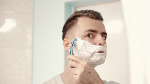Молодой человек бреется перед зеркалом в ванной комнате — стоковое видео