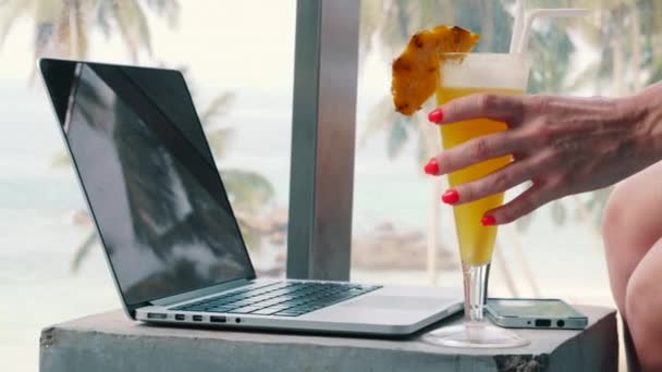 Jonge vrouw neemt een glas met een cocktail in de hand op de achtergrond van een laptop — Stockvideo