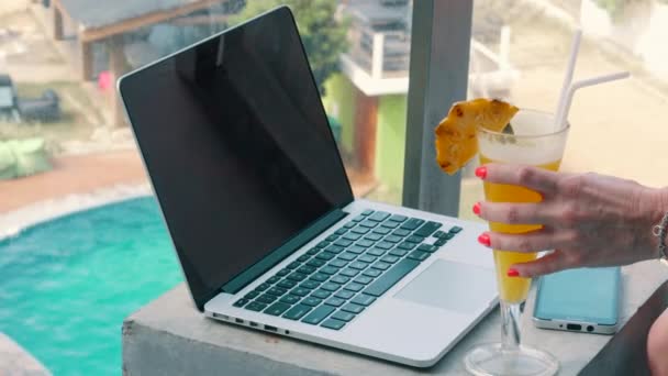 Молодая женщина берет в руки стакан с коктейлем на фоне ноутбука — стоковое видео